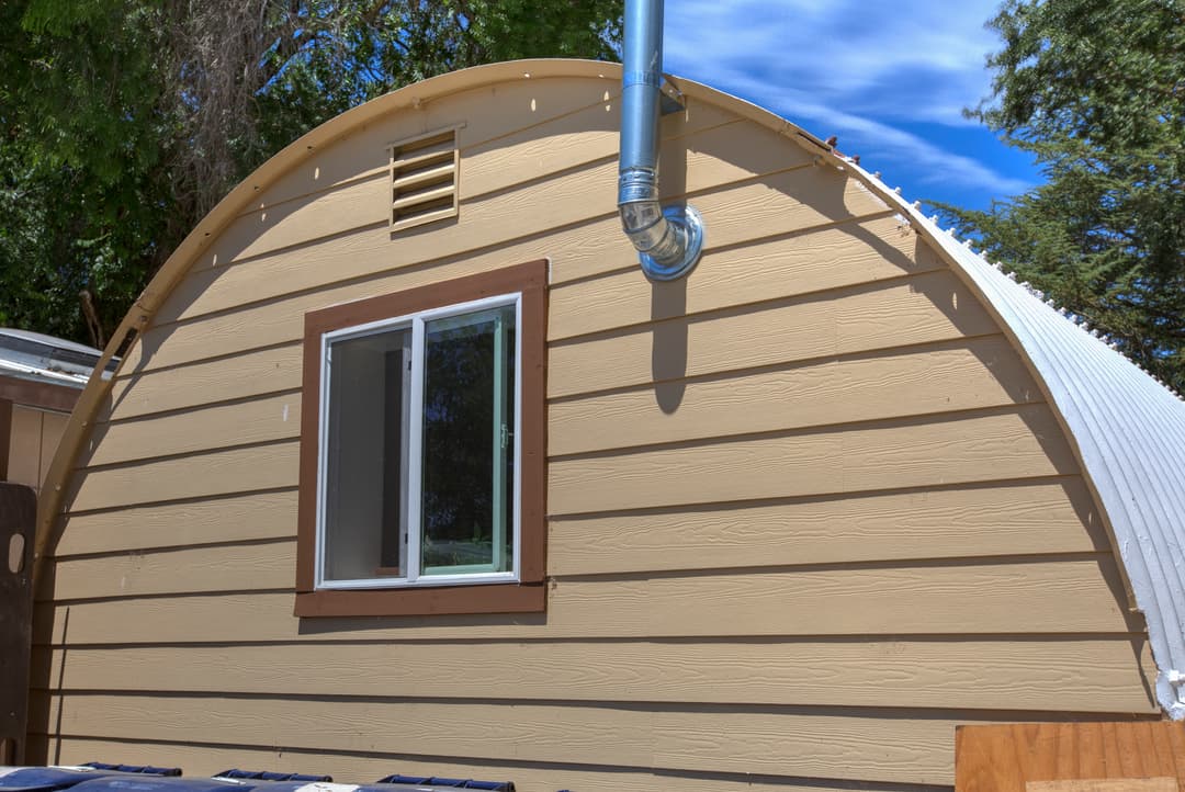 Exterior of arched tiny house with window, vent, and chimney pipe under blue sky