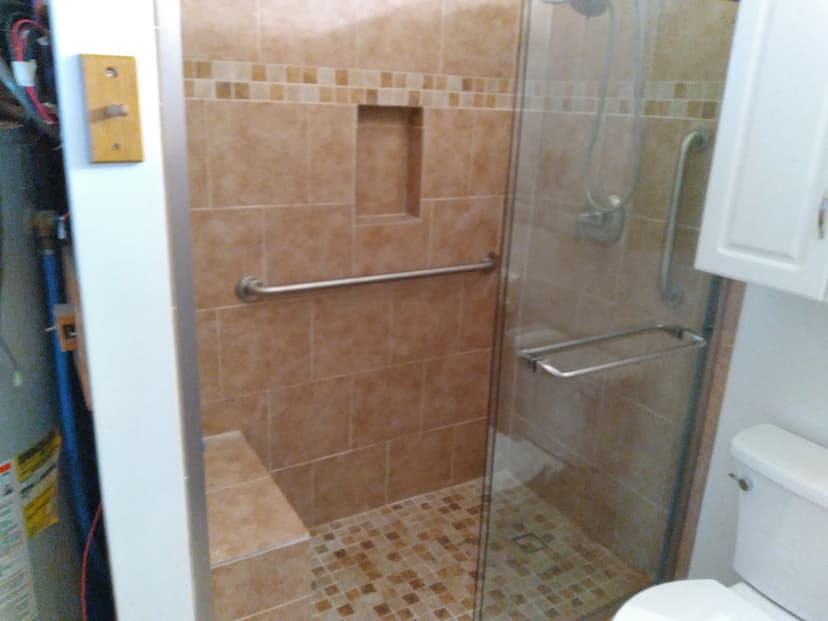 Modern walk-in shower with tan tiles, glass door, bench, and grab bars