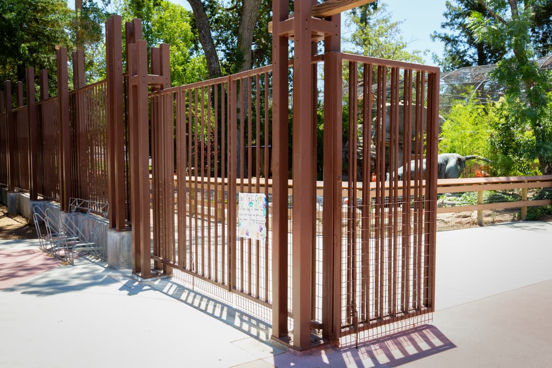 Brown metal zoo entrance gate under sunlight with trees and dinosaur sculptures in background